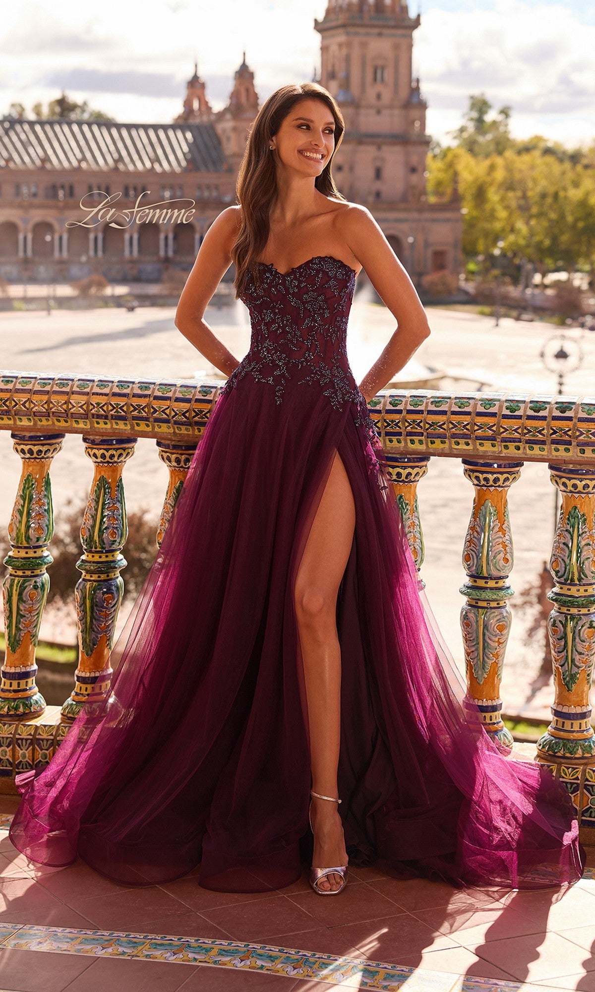 Wearing the Long Prom Dress: La Femme 33551, a woman in a strapless corset bodice and deep purple shade smiles on a tiled balcony, with historic buildings and trees behind her.